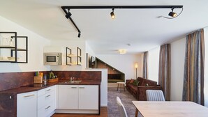 Una cocina moderna con armarios blancos y encimera oscura, un comedor con mesa y sillas, y una sala de estar con sofá y televisor.