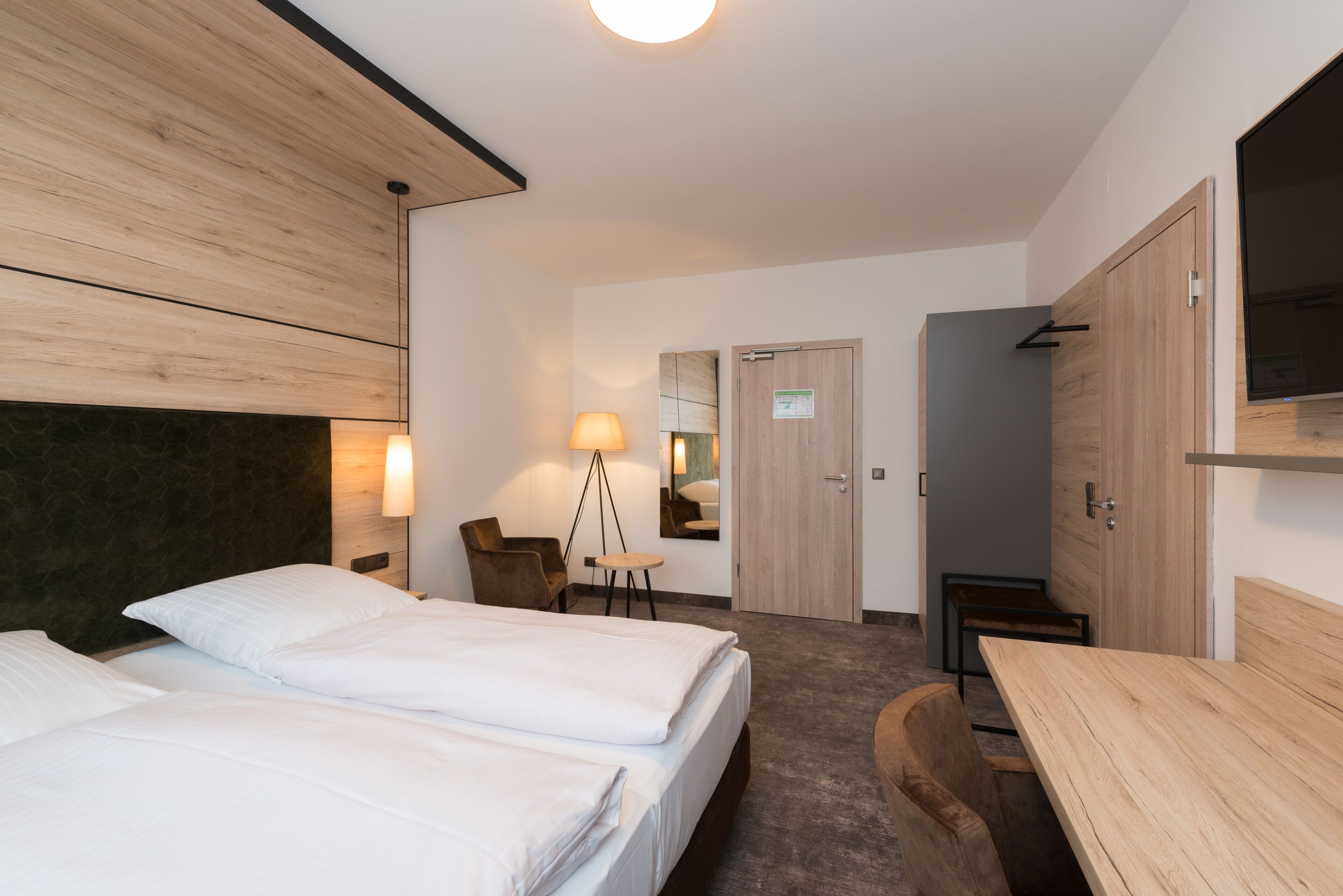A modern hotel room with two beds, a desk, a chair, a lamp, and a mirror.