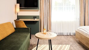 Habitación de hotel con sofá verde, una mesa de madera con una botella y vasos, un televisor montado en la pared y una cama.