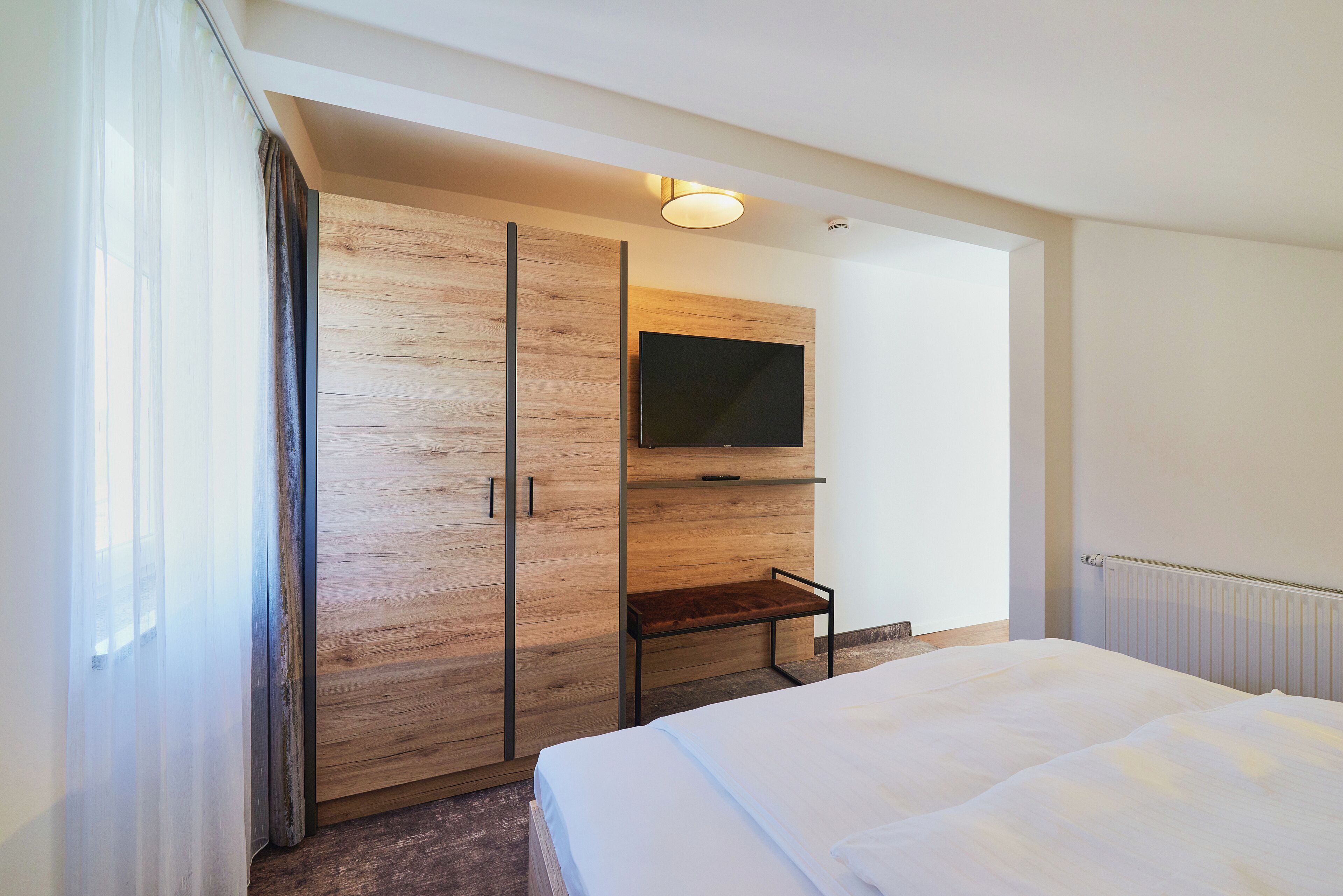 A modern hotel room with a large bed, a wooden wardrobe, a television mounted on the wall, and a small table.