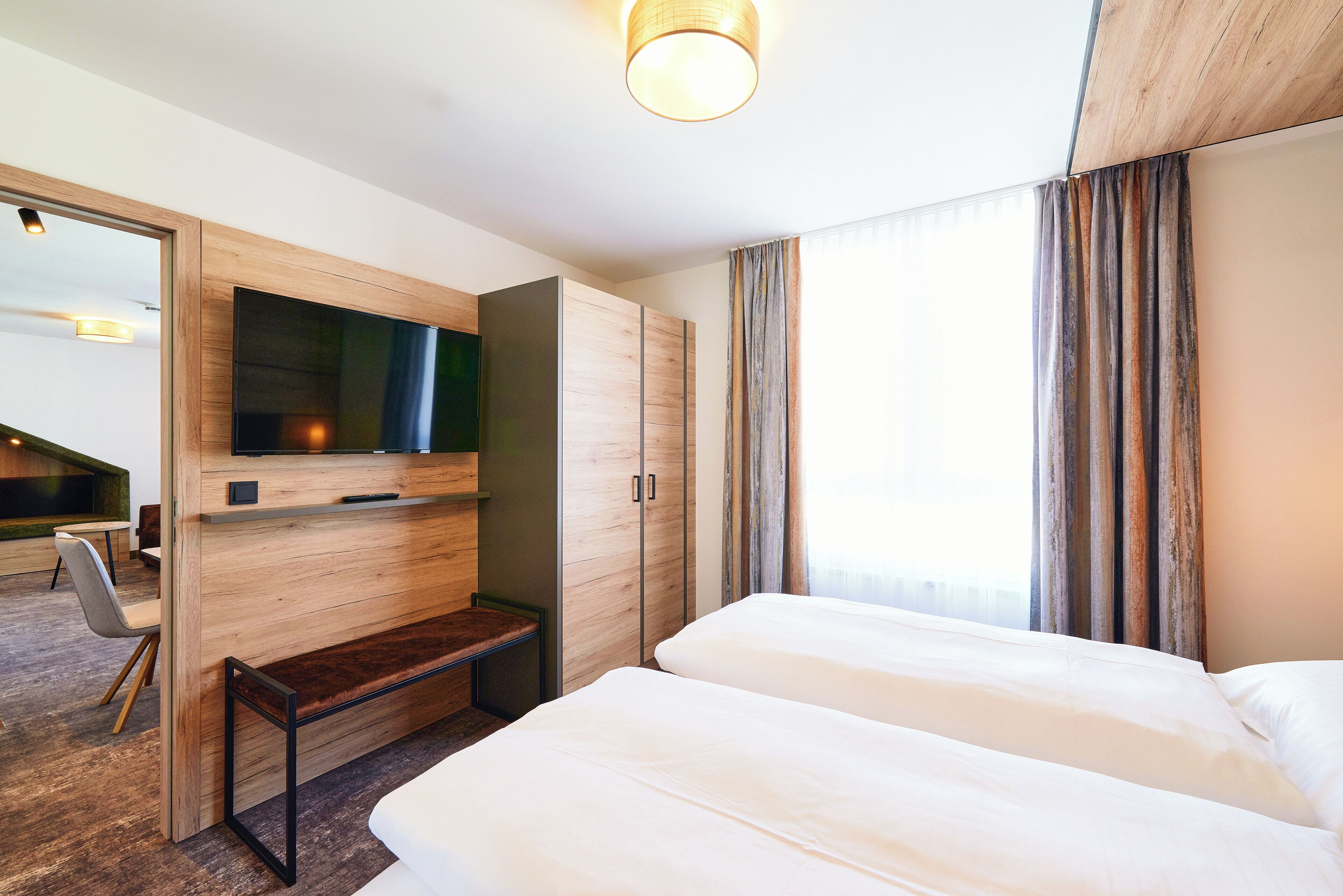 Een hotelkamer met twee bedden, een aan de muur bevestigde televisie, een houten garderobe en een bankje.