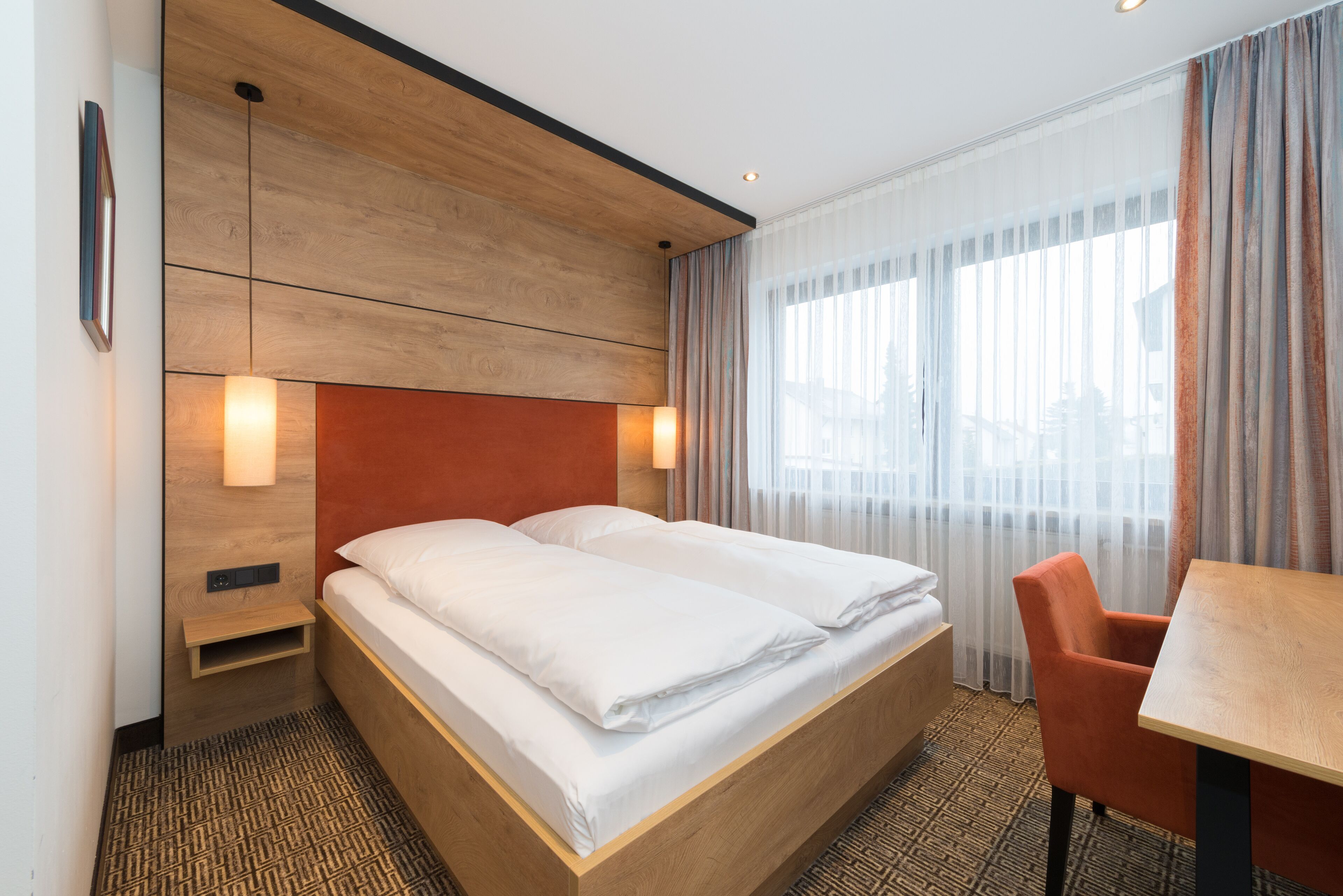 Una habitación de hotel con cabecera de madera, cama con ropa blanca, mesita de noche pequeña, una silla y un ventanal con cortinas traslúcidas.