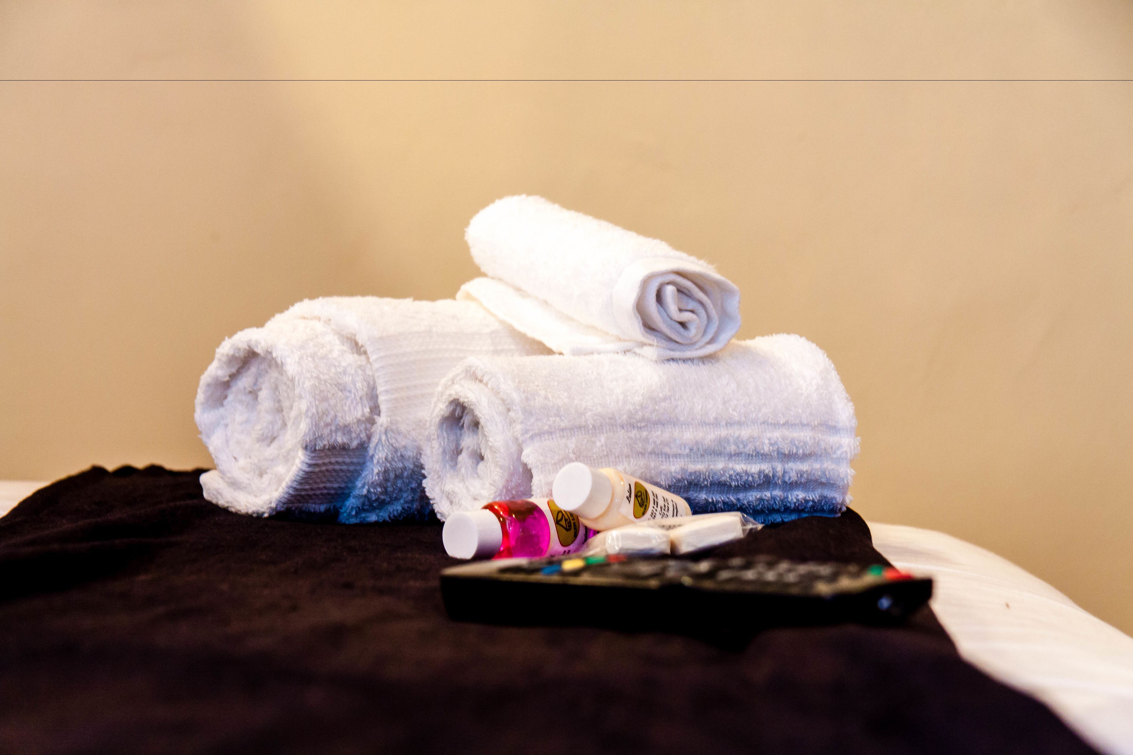 A bed with a black blanket, a rolled white towel, and a remote control.