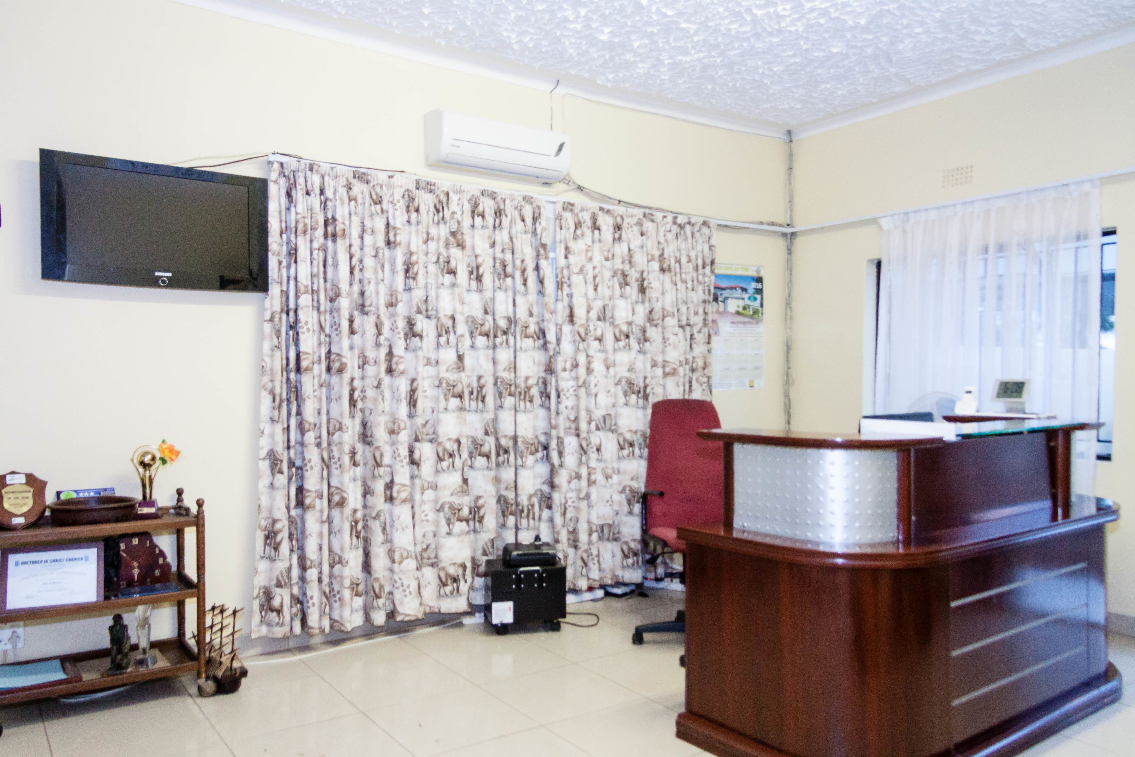 A reception area with a wooden desk, a red office chair, a flat-screen TV mounted on the wall, and patterned curtains.