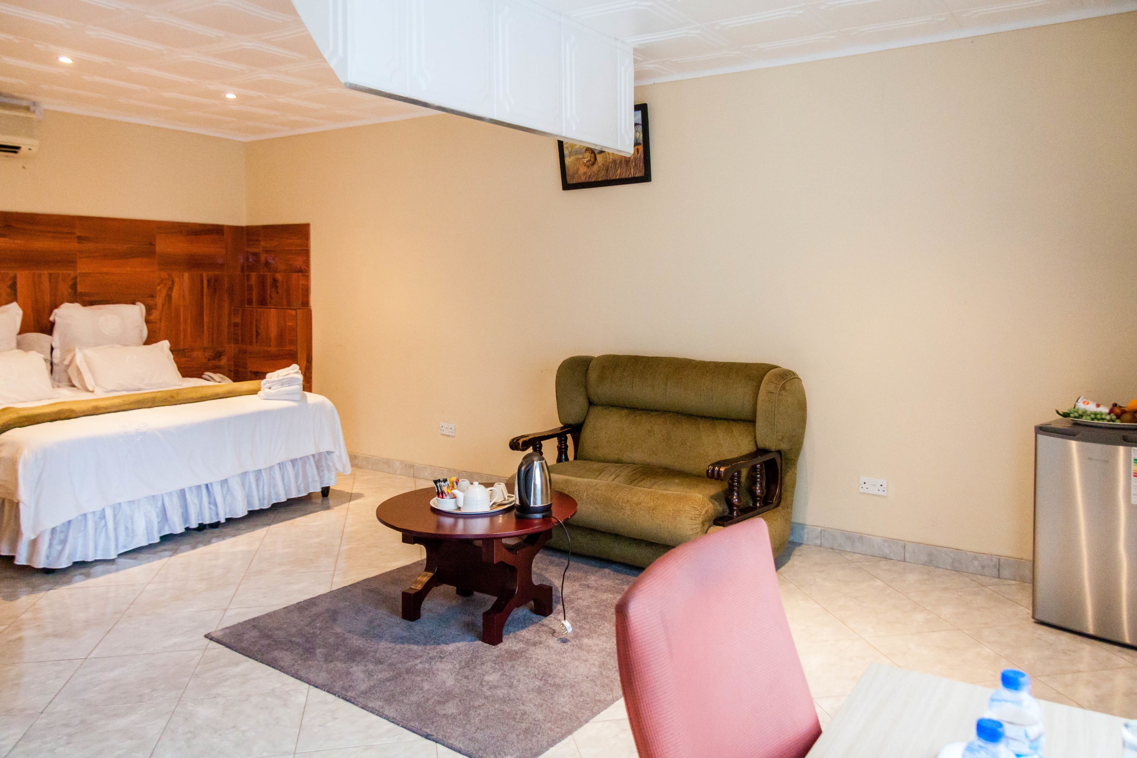 Une chambre d’hôtel avec un lit, un fauteuil vert, une petite table avec une bouilloire et un réfrigérateur.