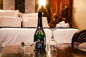 A bottle of JICROUX champagne and a wine glass on a wooden table in a hotel room with a bed and a nightstand with a lamp. - Glen Lodge & Tours (Bulawayo)