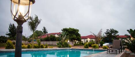 A swimming pool with a tiled edge, surrounded by palm trees and a building with a red roof in the background.