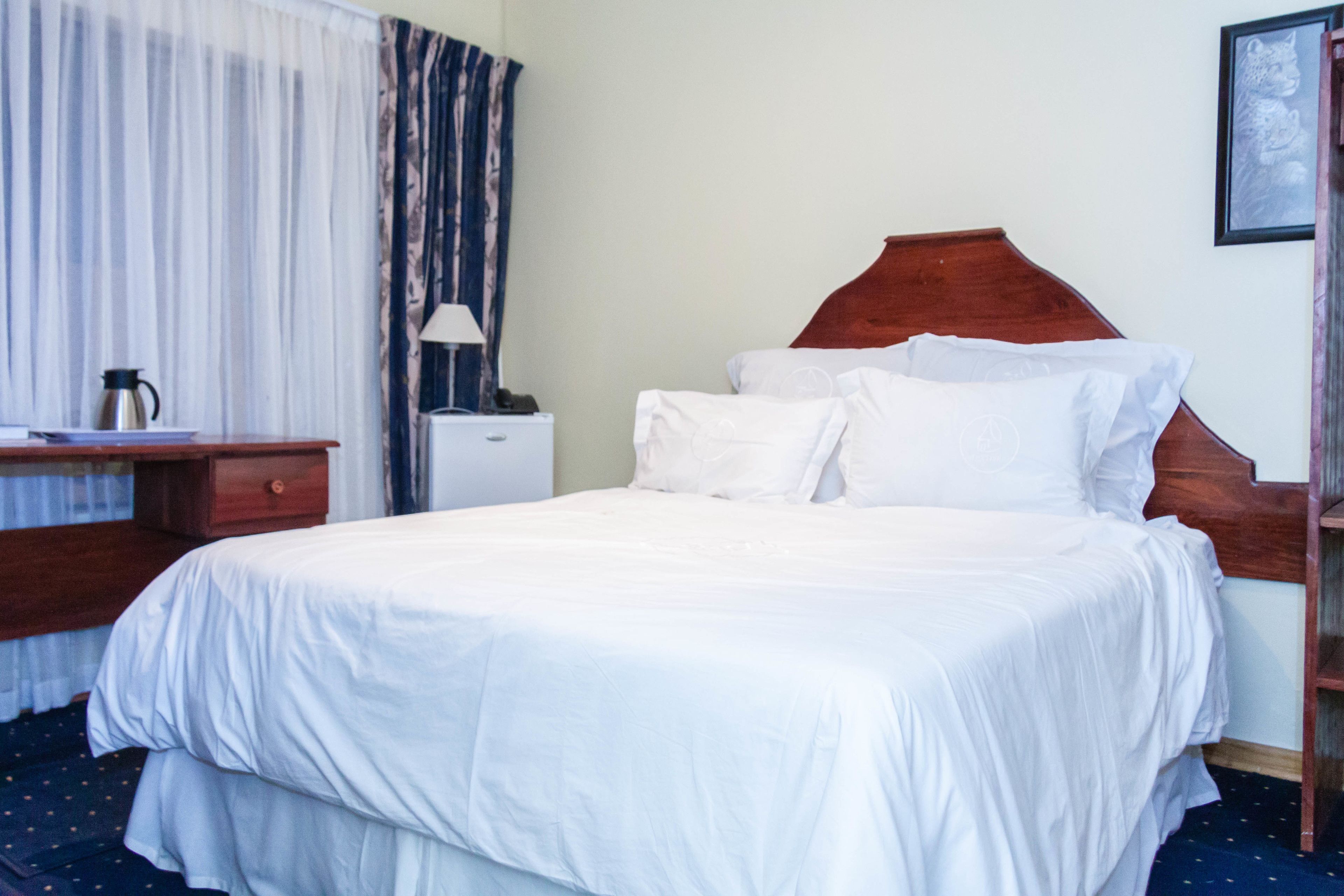 A neatly made bed with white linens, a wooden headboard, a bedside table with a lamp, and a small nightstand with a coffee pot.
