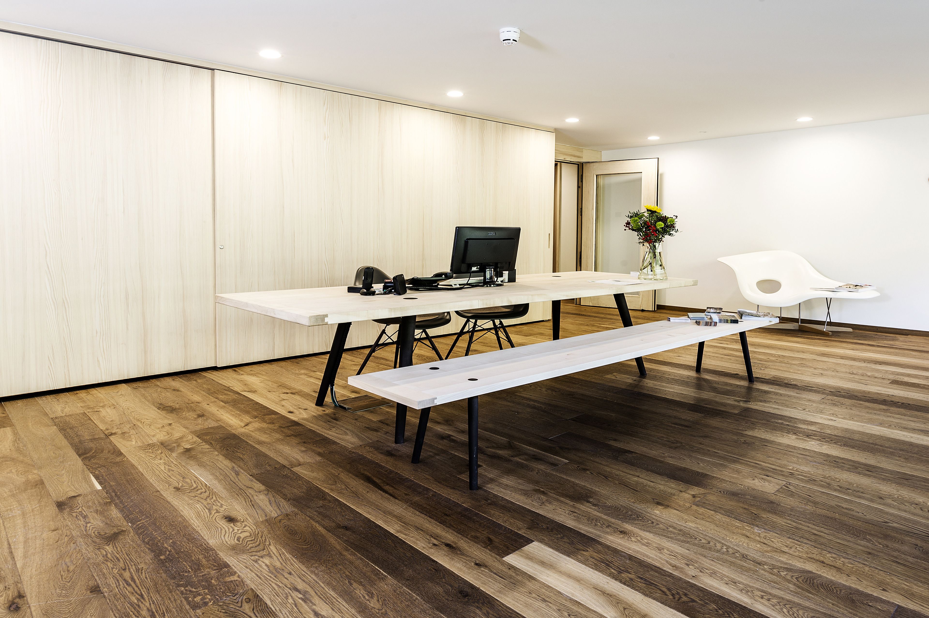A modern office space with a wooden floor, a white desk with a computer, a bench, and a white chair.