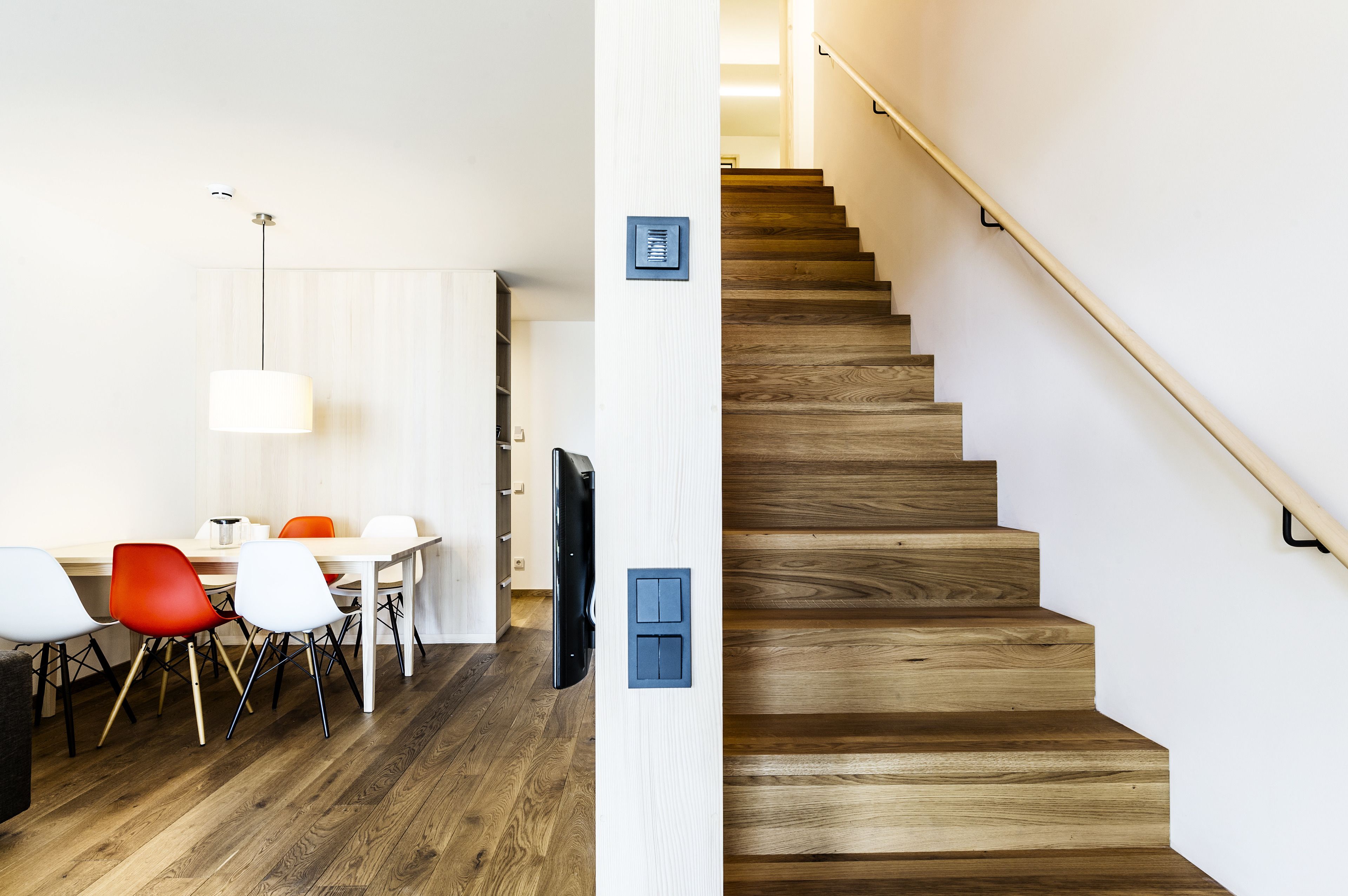 A modern interior with a dining area featuring a wooden table and chairs, a flat-screen TV, and a staircase with wooden steps and a metal handrail.