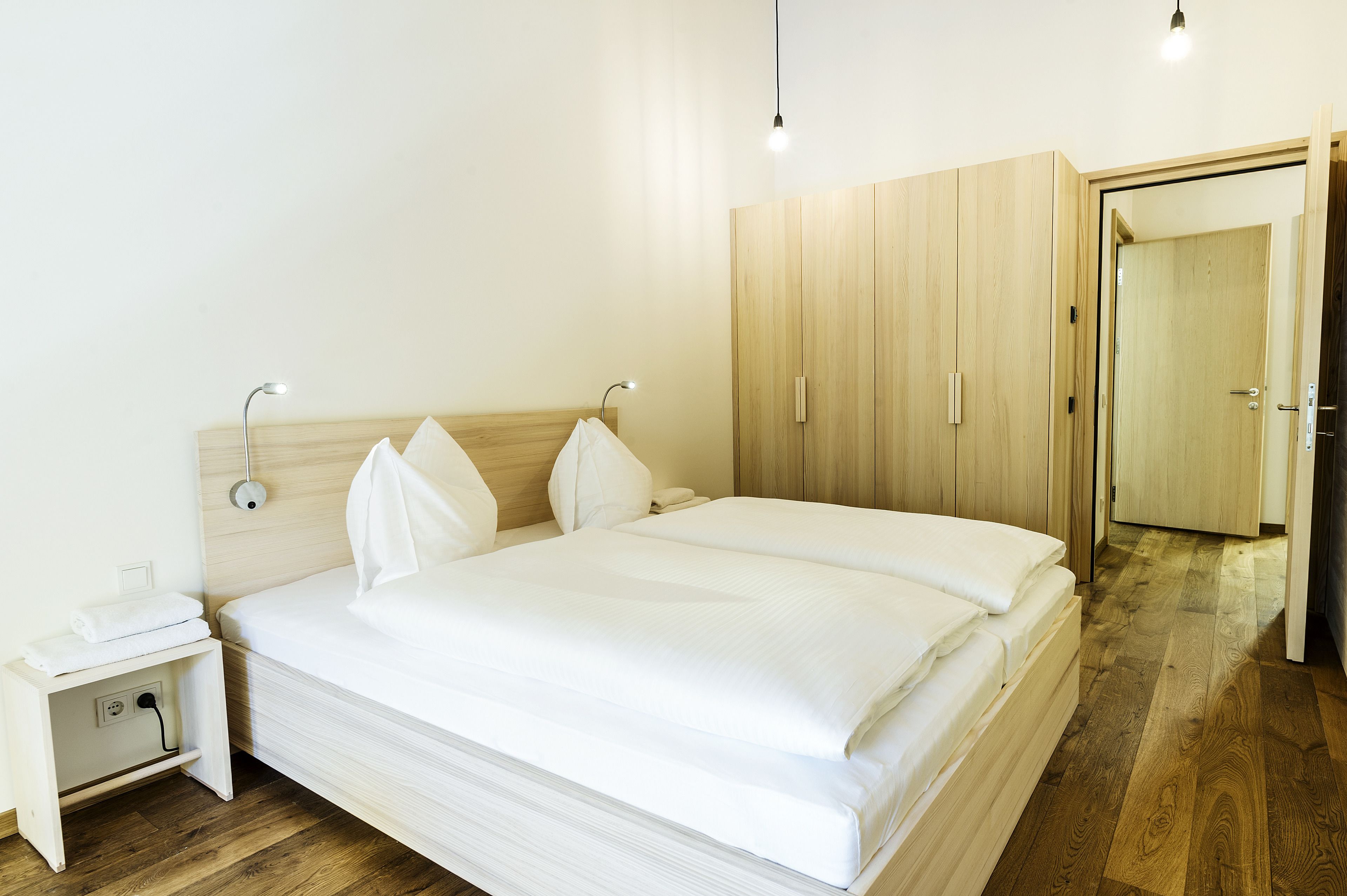 A neatly made bed with white linens, a bedside table with a lamp, and wooden wardrobe with a door.