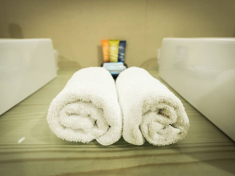 Two neatly rolled white towels placed on a bed with a beige headboard and a small box of toiletries in the background.