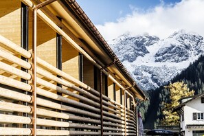 A building with a wooden facade and large windows, set against a backdrop of snow-capped mountains and a forested area. - alpen select apartments Kleinwalsertal (Mittelberg)