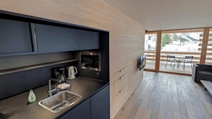 A modern kitchen with dark cabinets, a stainless steel sink, a coffee maker, and a microwave. There is a wooden dining area with a table and chairs visible through a glass door. - alpen select apartments Kleinwalsertal (Mittelberg)