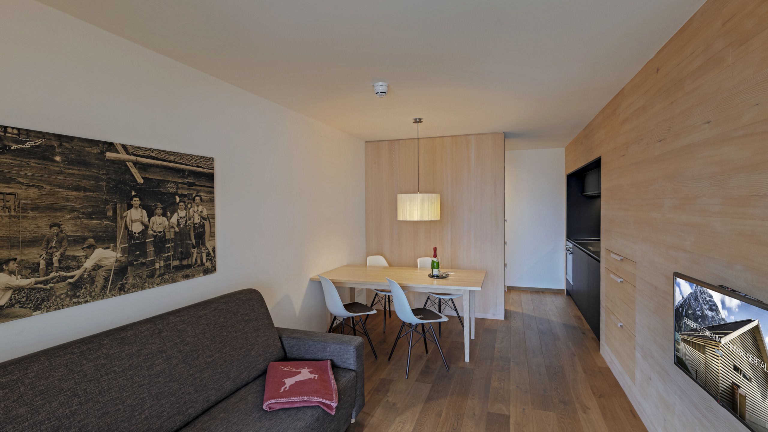 A modern living room with a wooden floor, a grey sofa, a red throw pillow, and a dining area with a table and chairs.