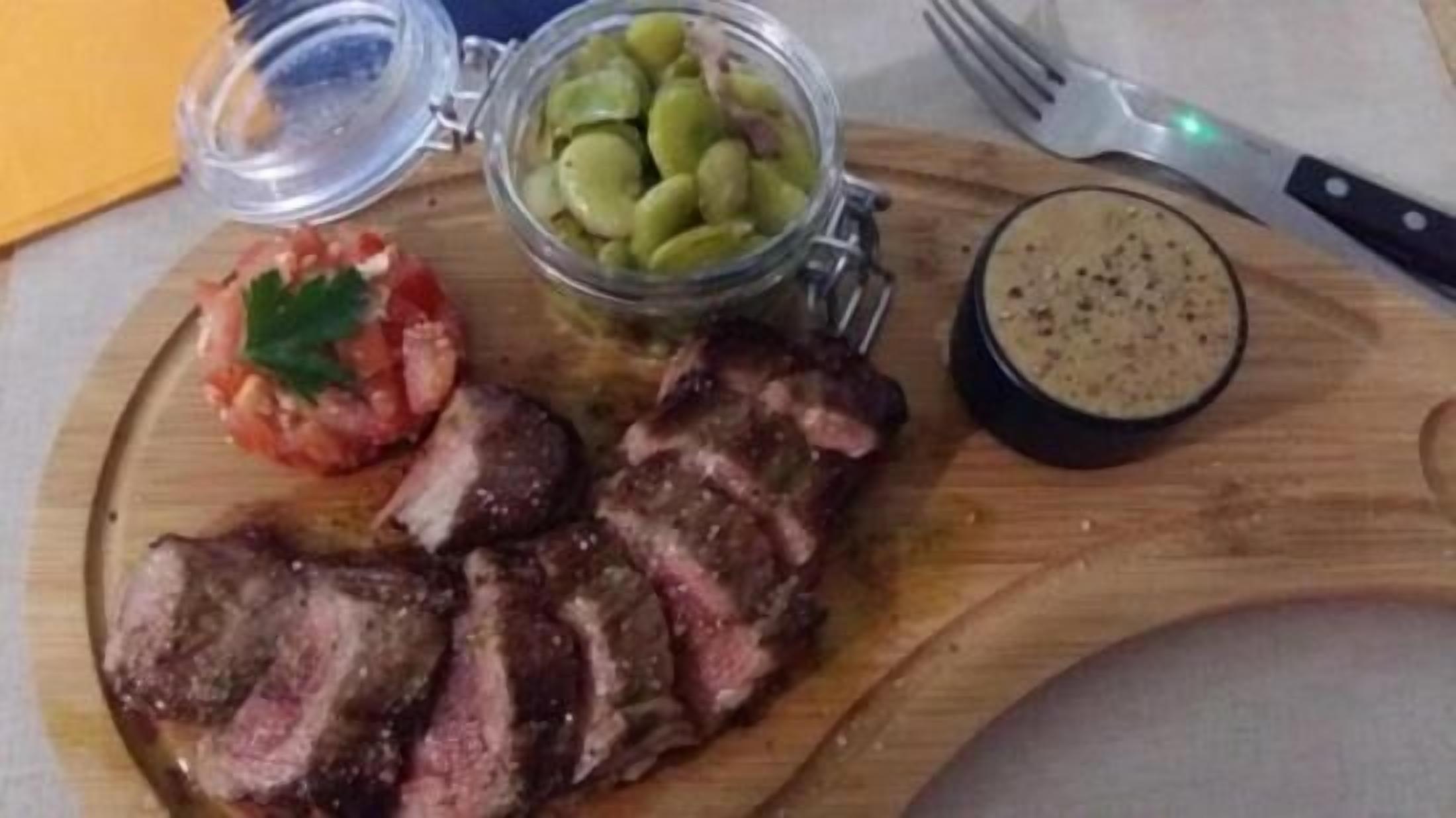 Une planche à découper en bois, avec un steak, un bocal de cornichons et un petit bol de sauce.