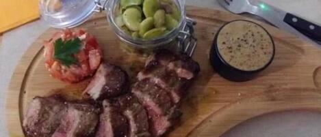 Une planche à découper en bois, avec un steak, un bocal de cornichons et un petit bol de sauce.