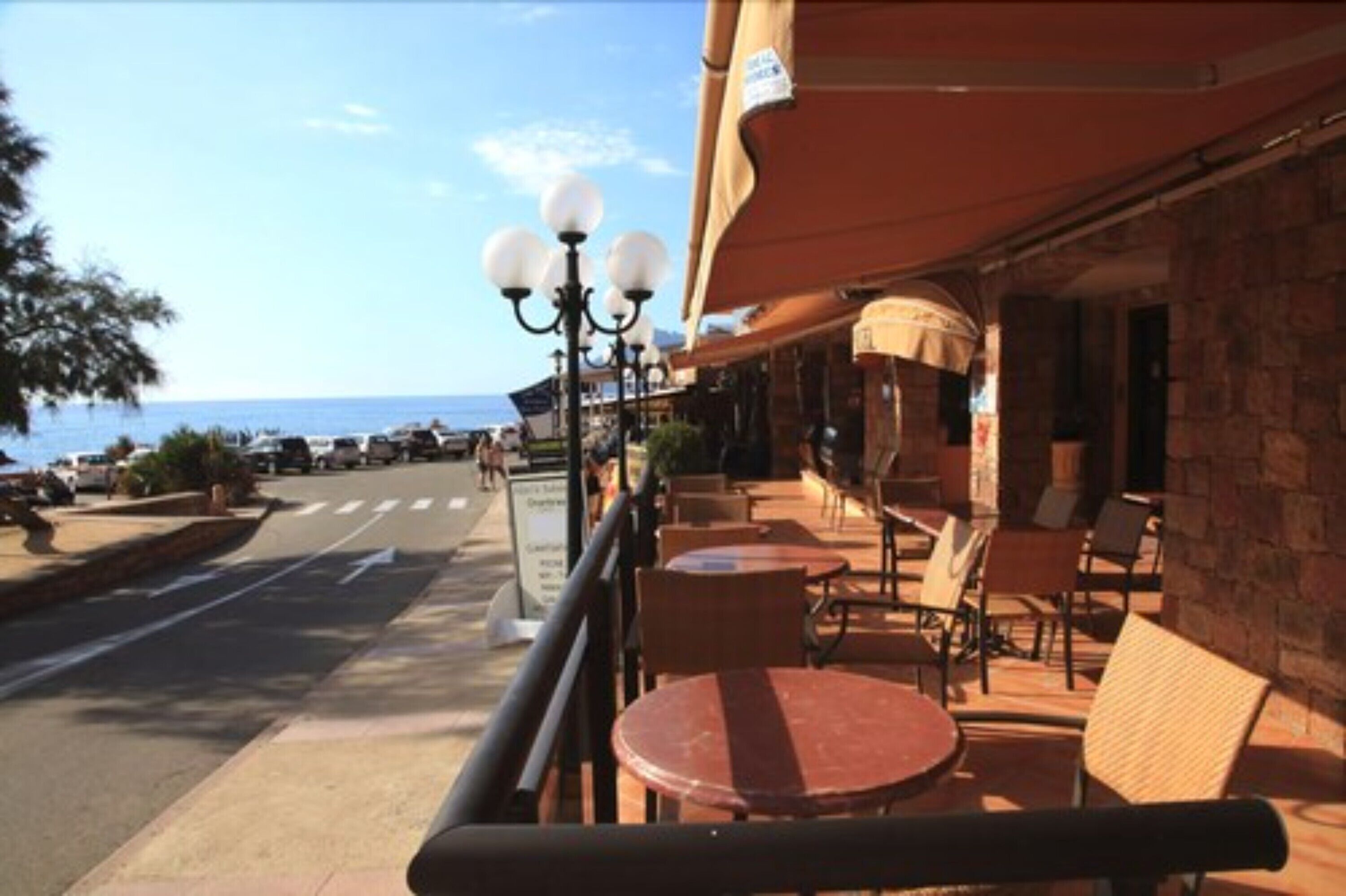 An outdoor seating area with tables and chairs, a street in the background, and a building with a stone facade.