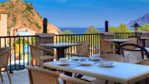 An outdoor dining area with wicker chairs and tables, overlooking a coastal landscape with a castle on a hill and the sea in the background. - Hôtel Idéal (Ota)