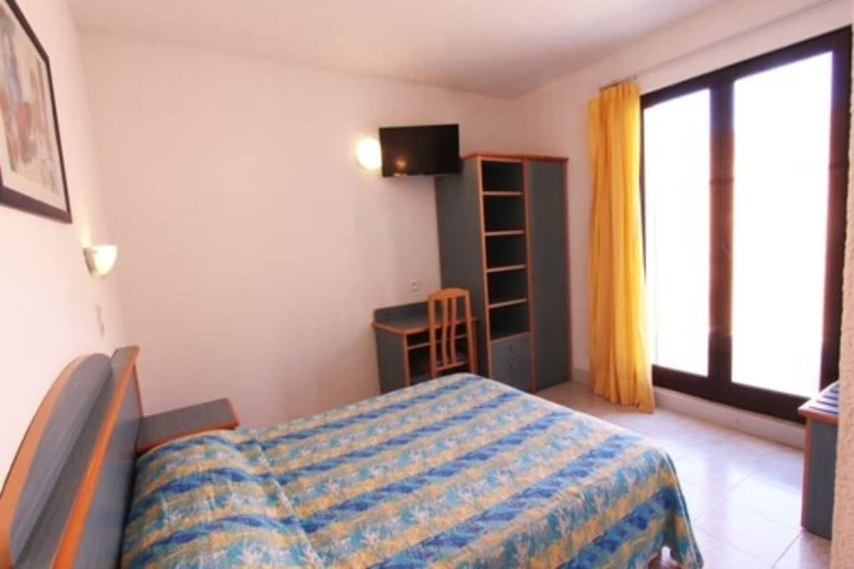 A bedroom with a bed, a desk, a chair, a TV, and a window with curtains.