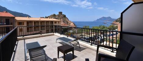 A balcony with outdoor furniture, a view of the sea, and buildings in the background.