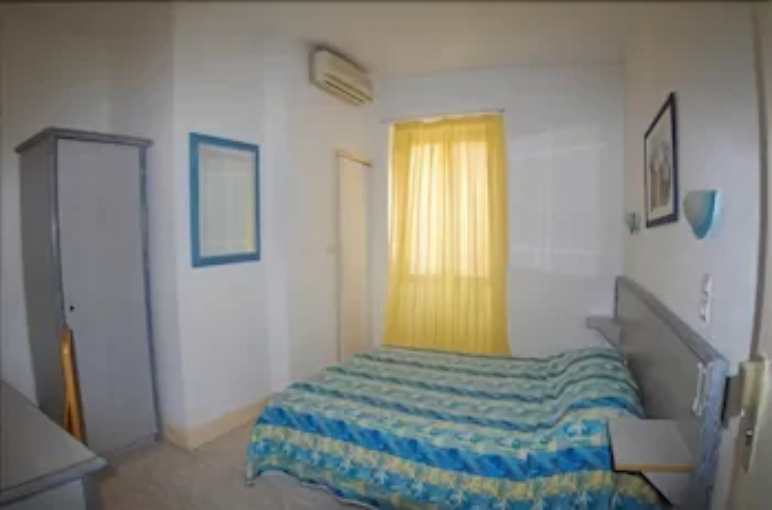 A bedroom with a bed, a wardrobe, a window with yellow curtains, and a wall-mounted air conditioner.