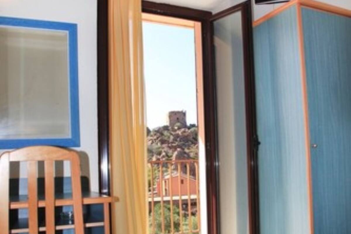 A room with a bed, a desk with a chair, a television, and a sliding door leading to a balcony with a view of a building.