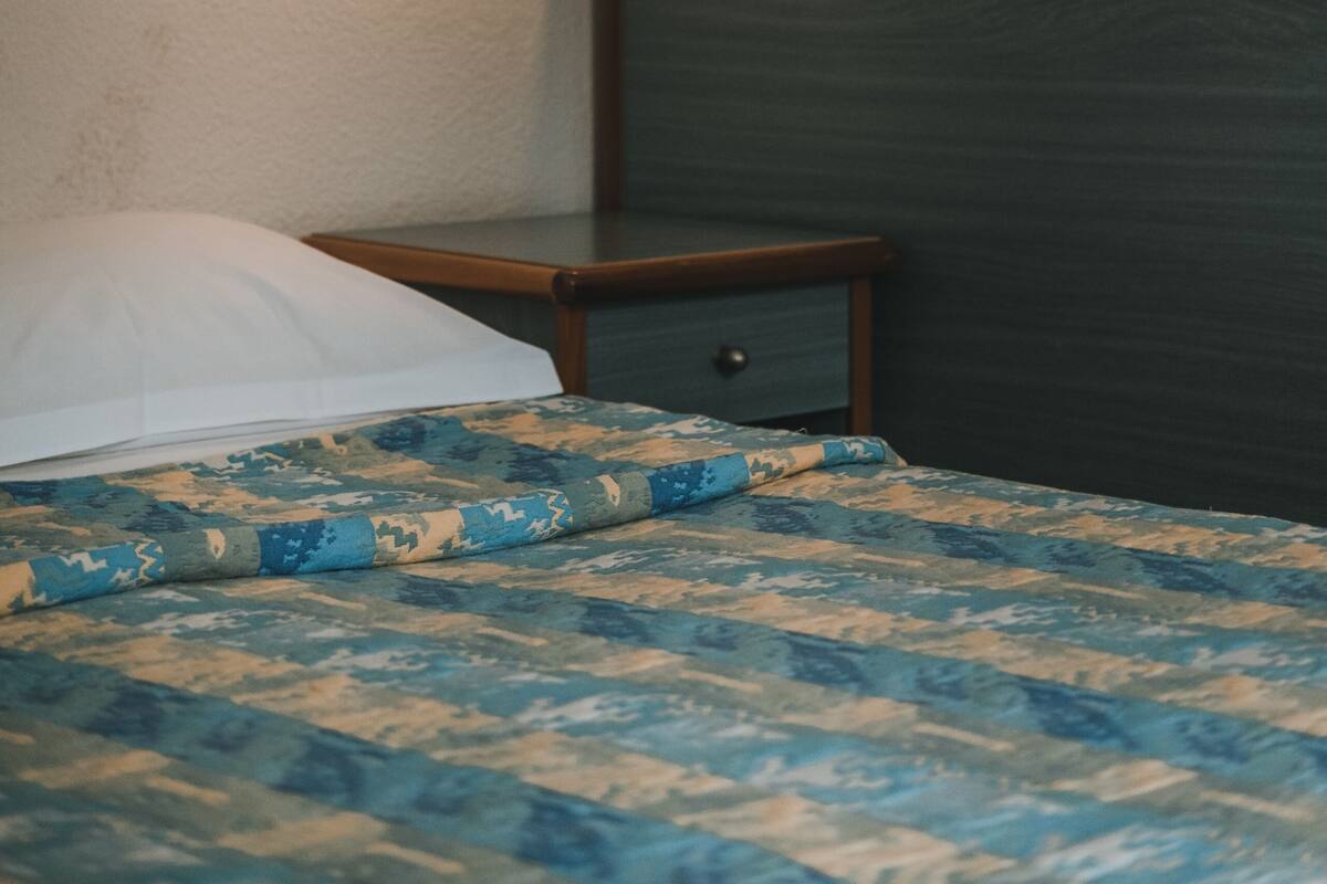 A neatly made bed with a patterned bedspread, a bedside table with a lamp, and a framed picture on the wall.