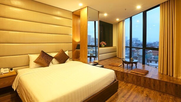 Una habitación de hotel moderna con una cama grande, una mesita de noche de madera, una pequeña mesa y un ventanal con vistas a la ciudad.