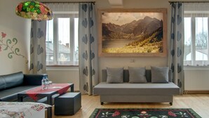 Ein modernes Wohnzimmer mit einem grauen Sofa, einem roten Tisch mit Blumenmuster-Tischdecke, einer Hängelampe und einem großen Gemälde von einer Berglandschaft an der Wand.