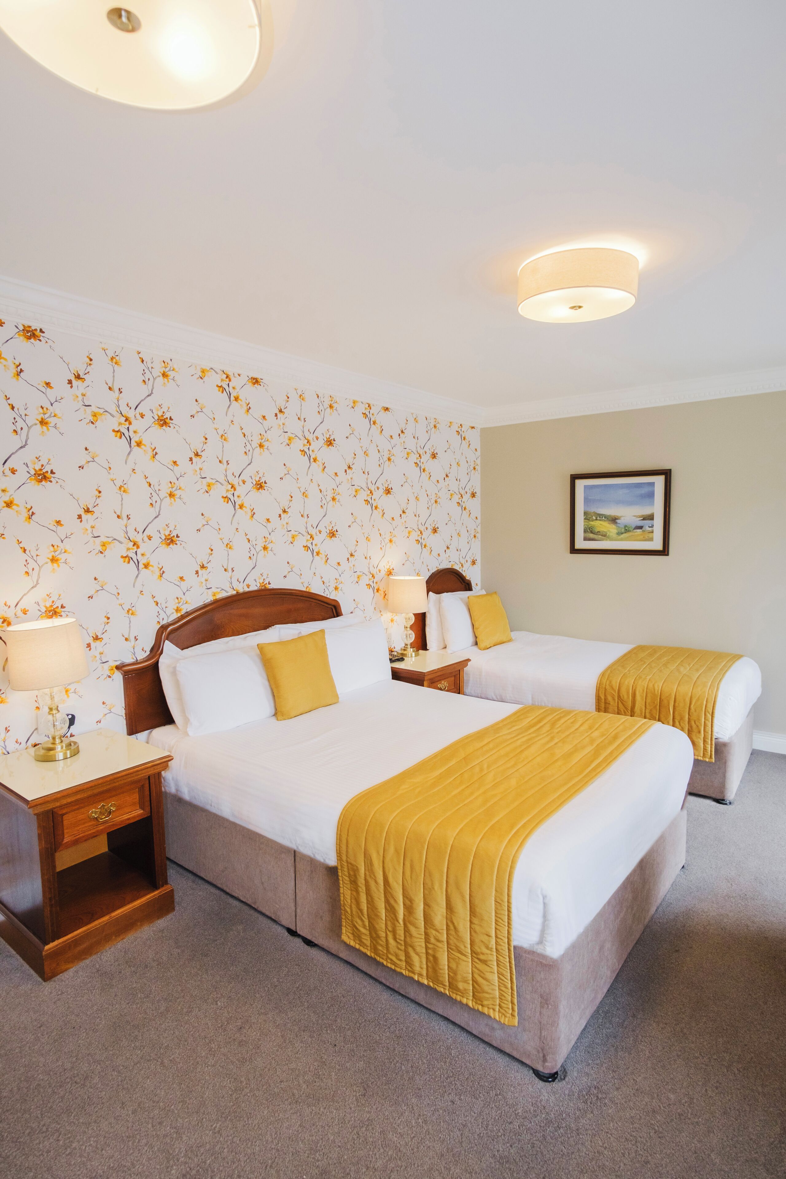Habitación de hotel con dos camas, una mesita de noche, una lámpara, papel tapiz floral y un cuadro en la pared.