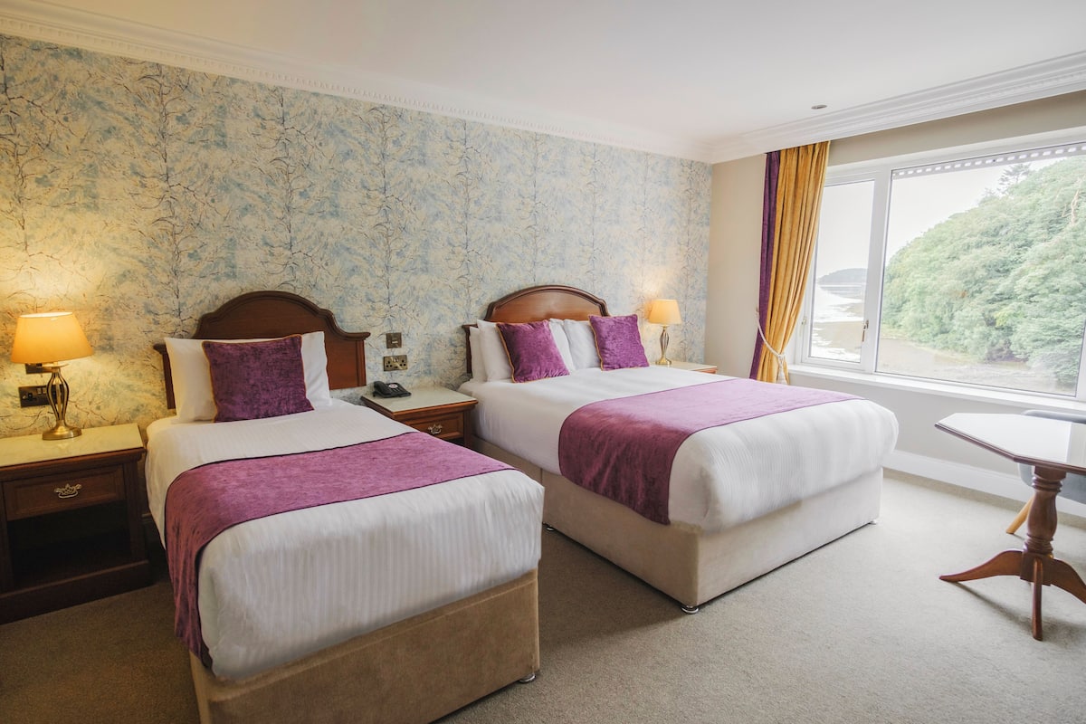A hotel room with two beds, a nightstand with a lamp, a small table, and a window with a view of greenery.