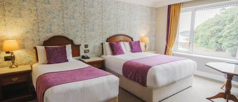 A hotel room with two beds, a nightstand with a lamp, a small table, and a window with a view of greenery.