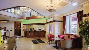 Un hall de hotel con mostrador de recepción, una escalera, un sofá rojo, una mesa de comedor y una araña.