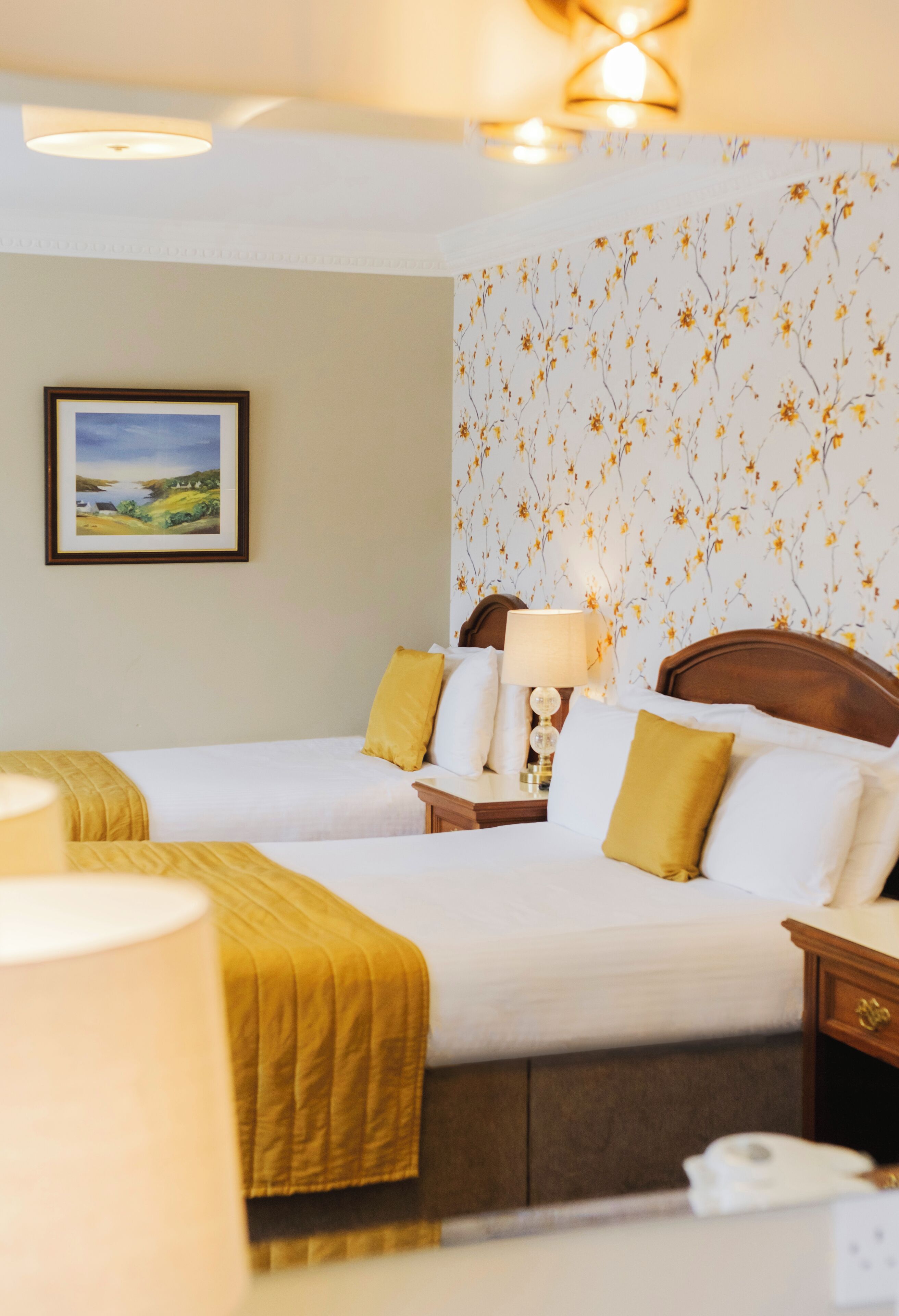 A hotel room with two beds, a nightstand with a lamp, a framed picture on the wall, and a floral wallpaper pattern.