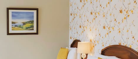 Ein Hotelzimmer mit zwei Betten, einem Nachttisch mit Lampe, einem gerahmten Bild an der Wand und einem floralen Wandpapier.