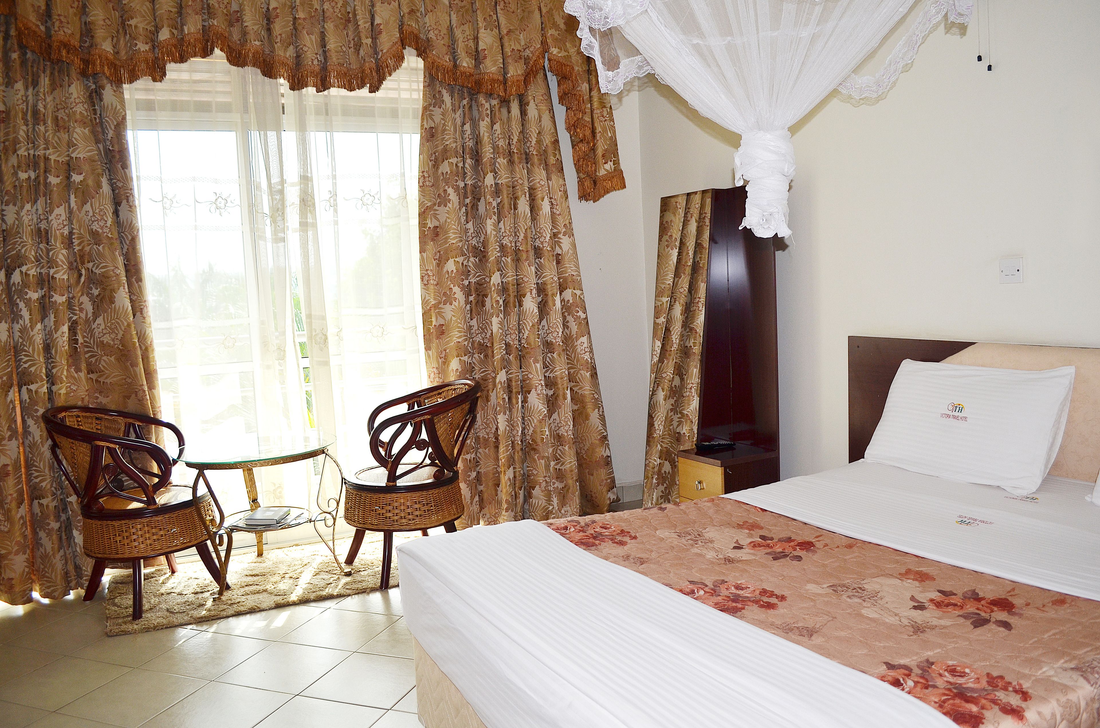 A hotel room with a bed, two wicker chairs, a small table, and a window with curtains.