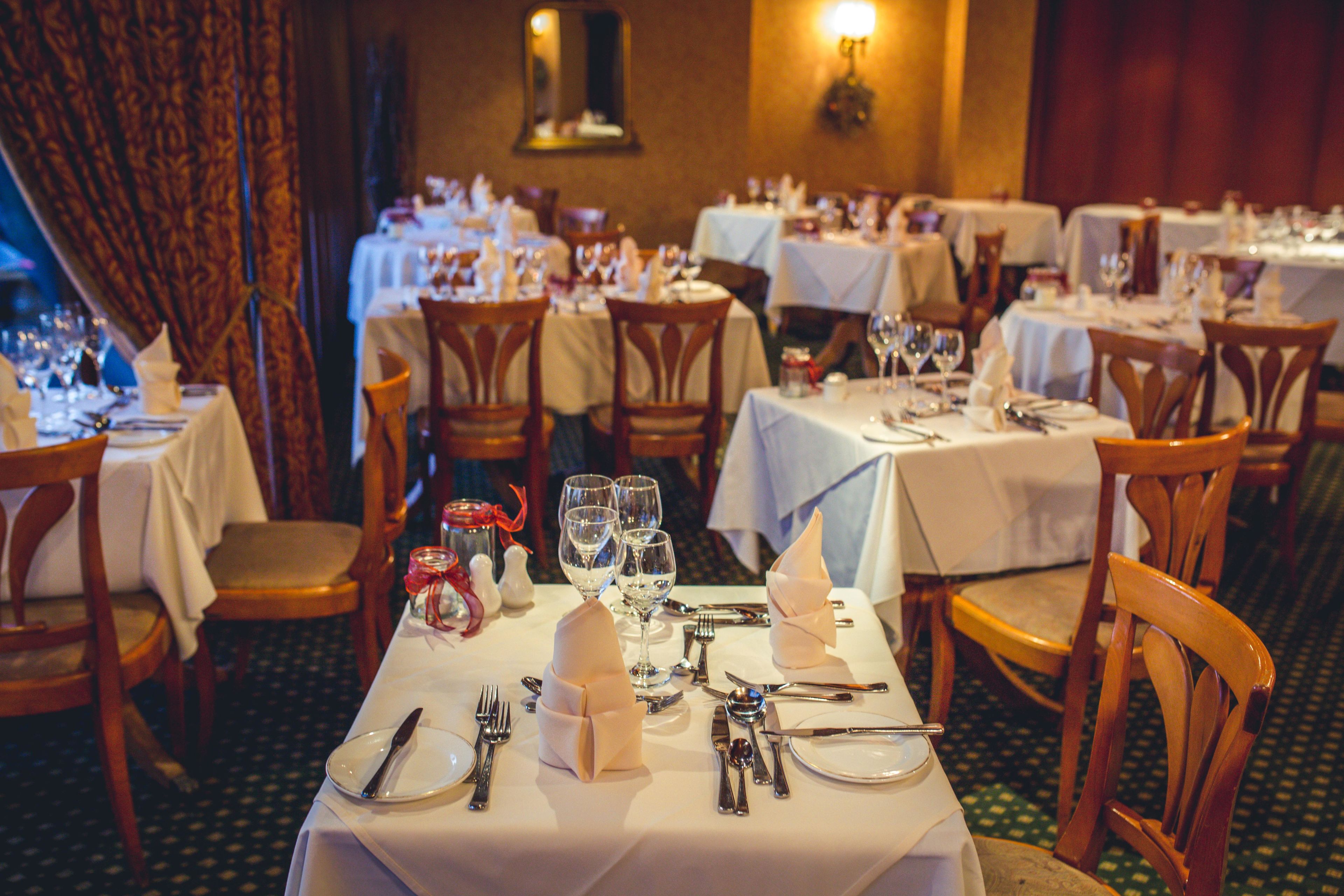 Un restaurante con varias mesas dispuestas para comer, cada una con manteles blancos, sillas de madera y cubertería ordenada.