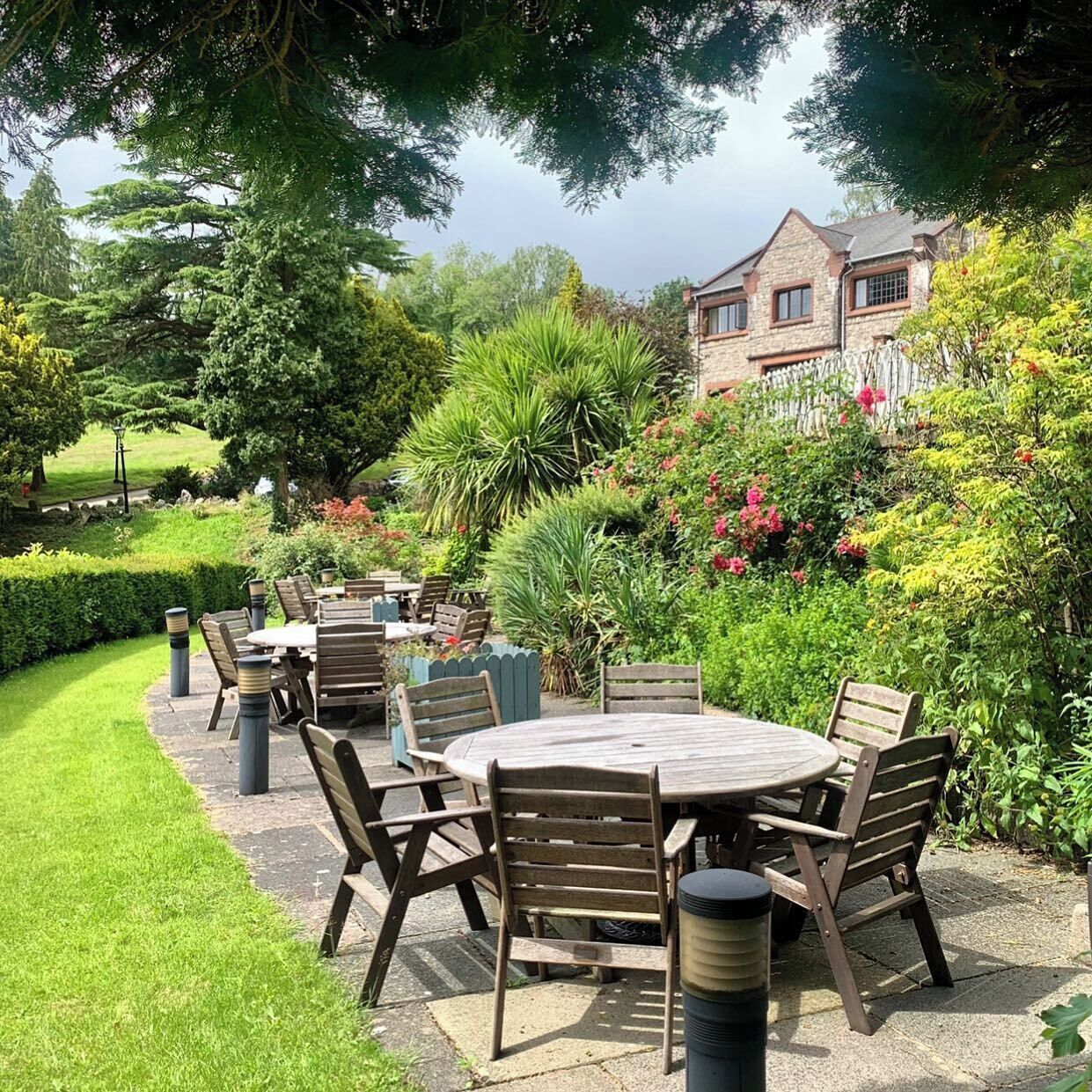 Une terrasse extérieure avec des tables rondes et des chaises en bois, entourée d’une végétation luxuriante et, au fond, d’un bâtiment en pierre.