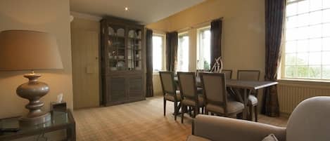 A dining area with a table and chairs, a lamp on a side table, and a view through a window.