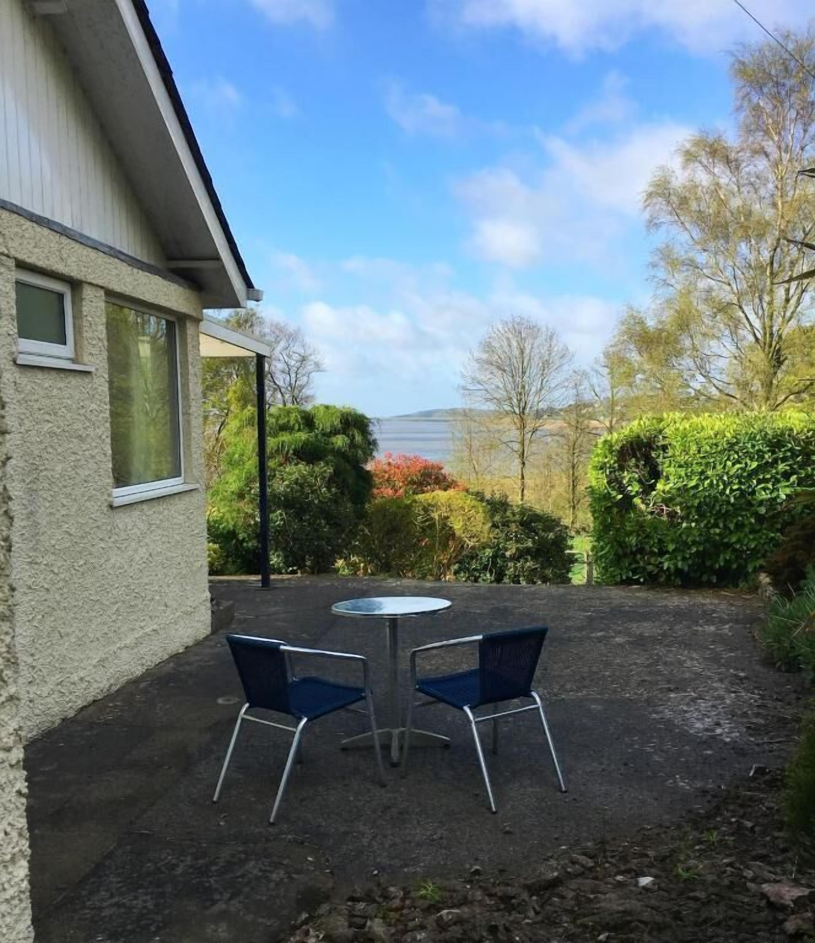 A patio with two chairs and a table, a building with a window, and a view of a body of water and trees.