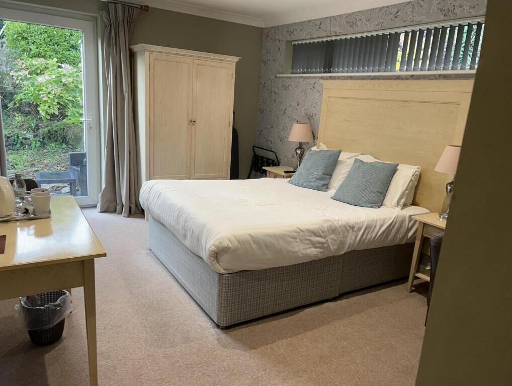 A bedroom with a large bed, a wooden headboard, a nightstand with a lamp, a window with blinds, and a view of greenery outside.