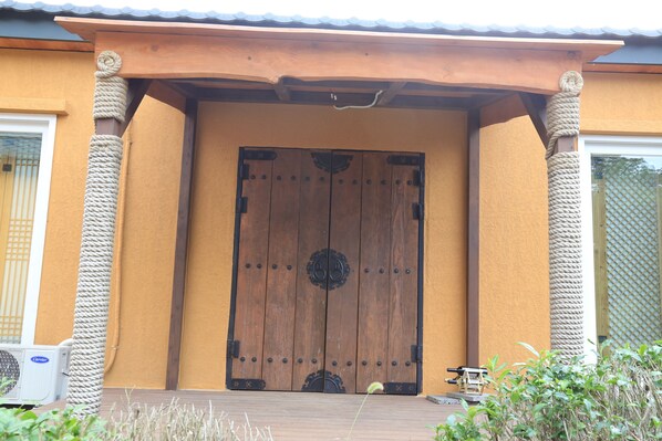 A large wooden door with a decorative metal handle, flanked by rope columns, and a small air conditioning unit to the left. - Jeju Best Hill (Jeju City)