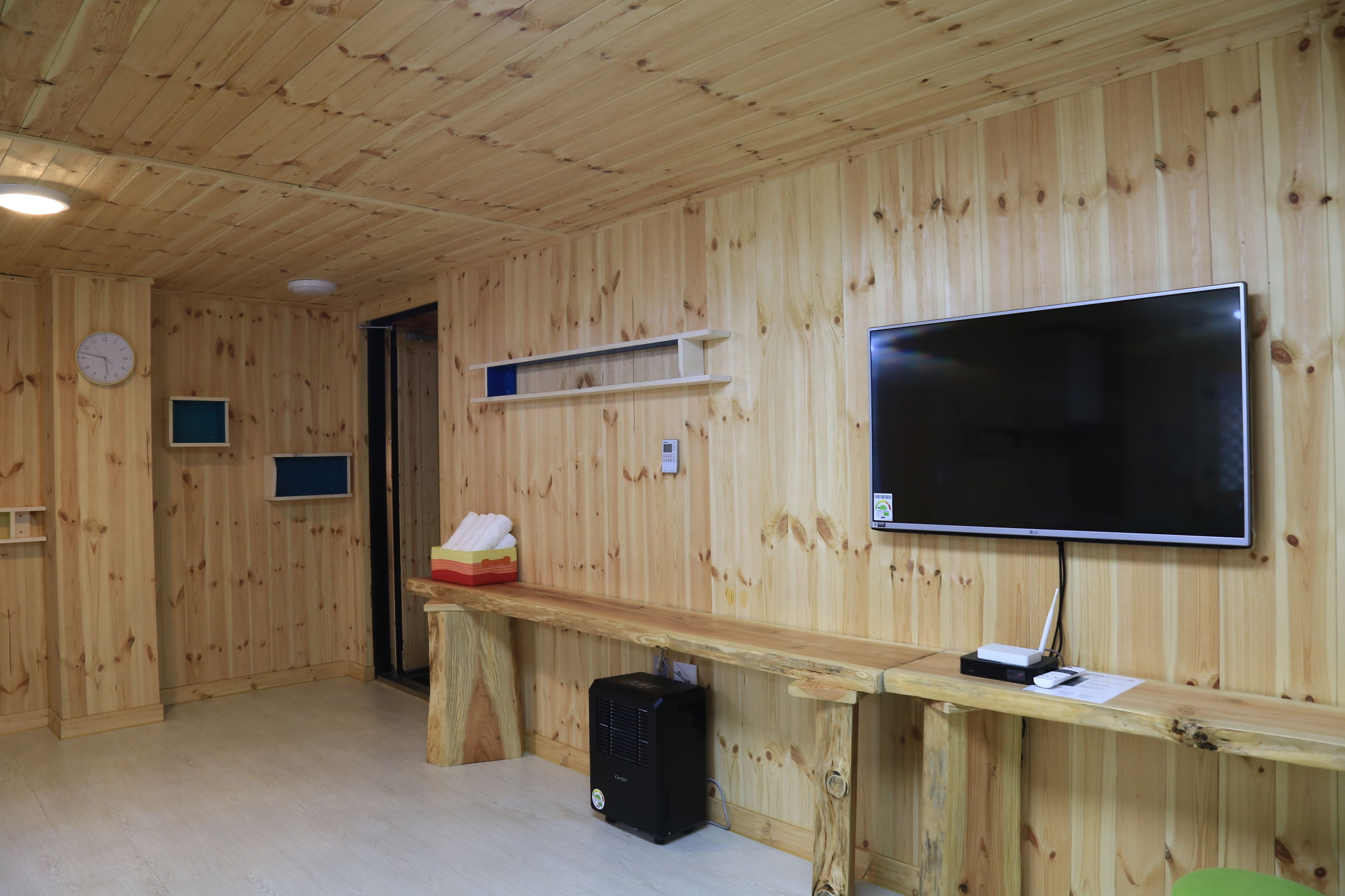 Una habitación con paneles de madera, un televisor de pantalla plana montado en la pared, un escritorio de madera con una computadora, y un estante pequeño con un reloj y una caja de pañuelos.