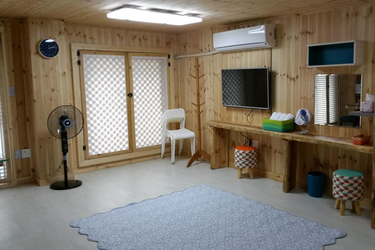 Een kamer met houten wanden, een televisie, een ventilator, een stoel, een bureau met een computer en een klein krukje.