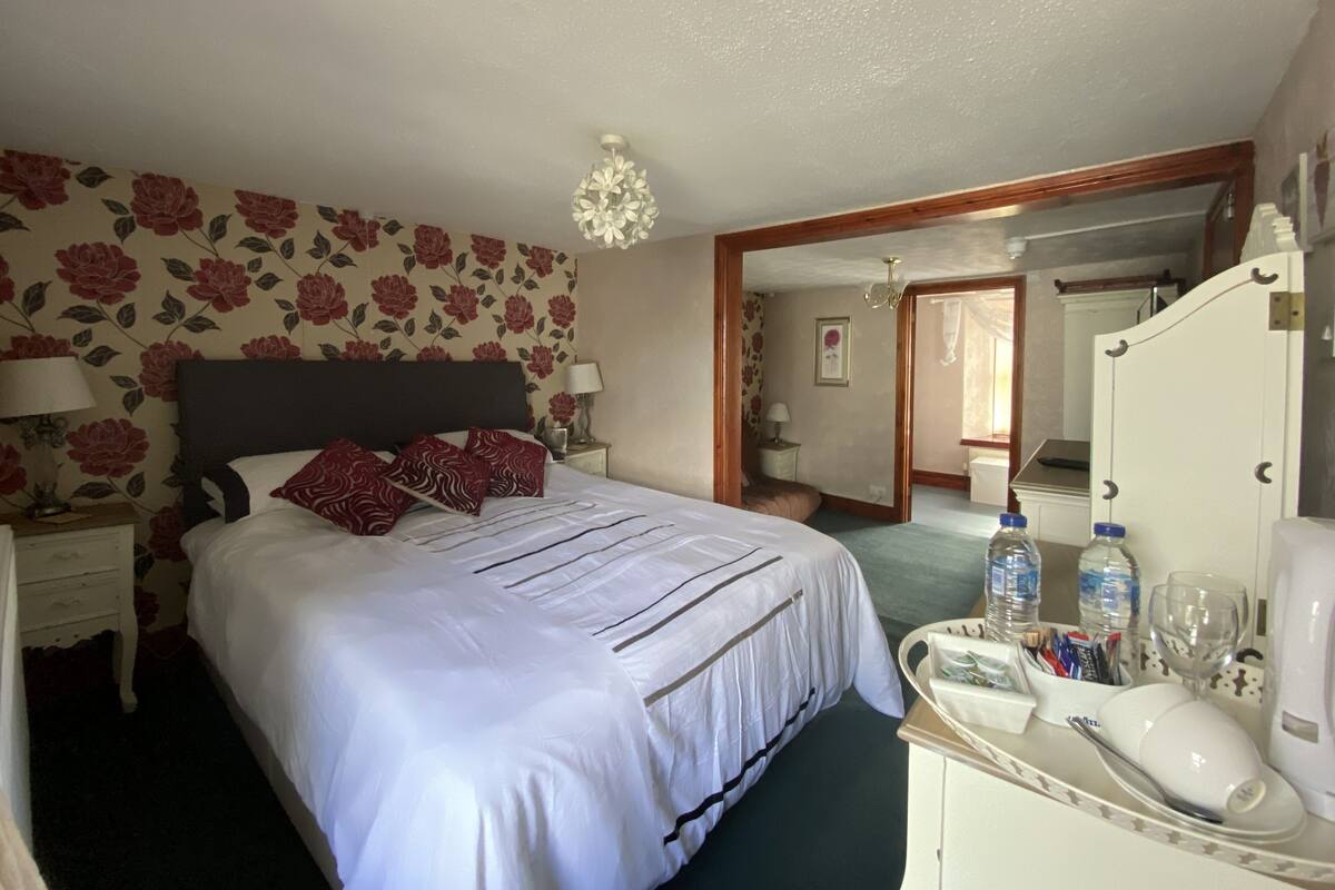 A hotel room with a bed, a nightstand, a dresser, a mirror, a chandelier, and a bathroom visible through an open door.
