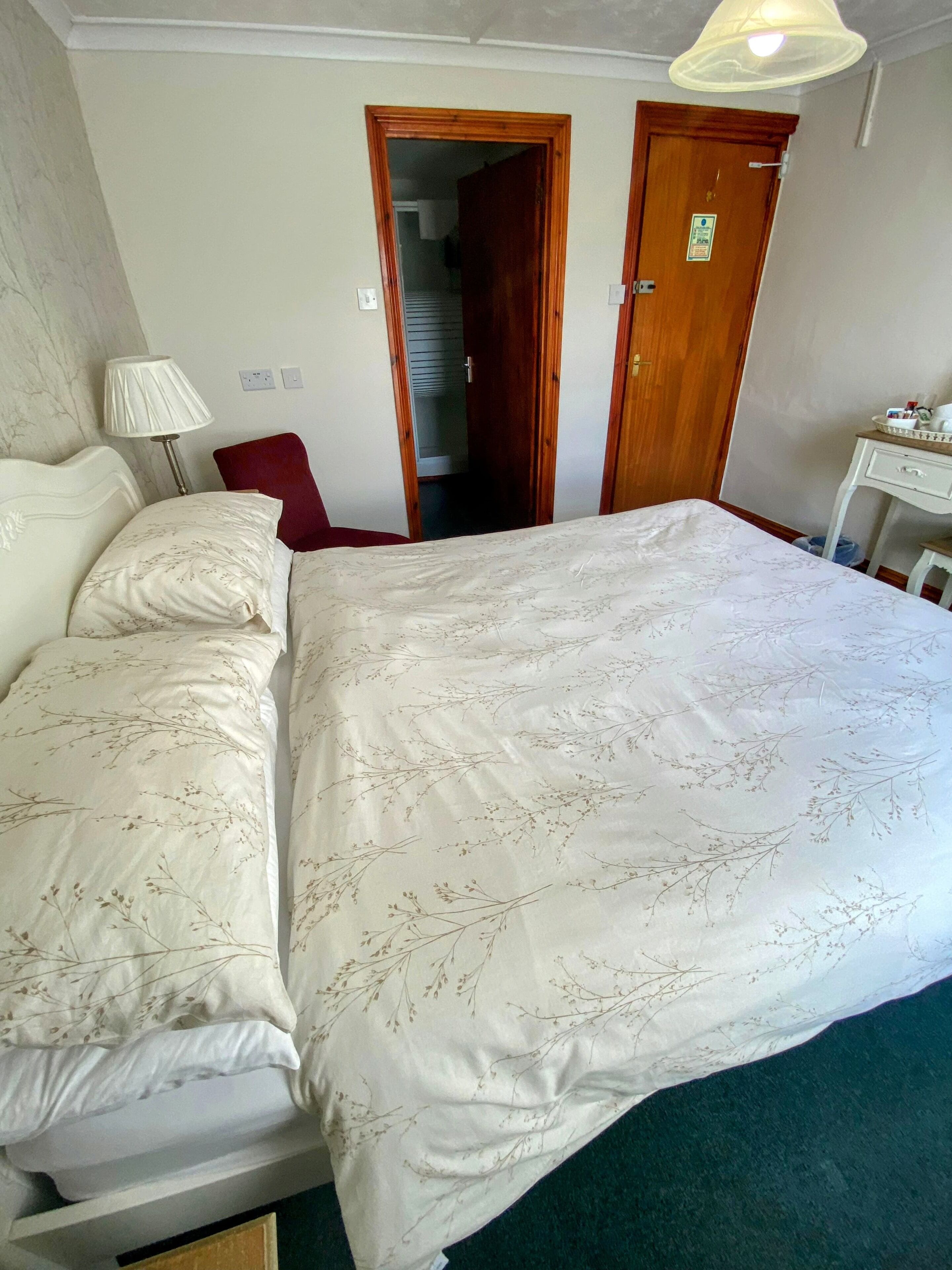 Cama bem arrumada com colcha floral, abajur e uma cadeira vermelha em quarto com porta de madeira e pequena mesa com objetos sobre ela.