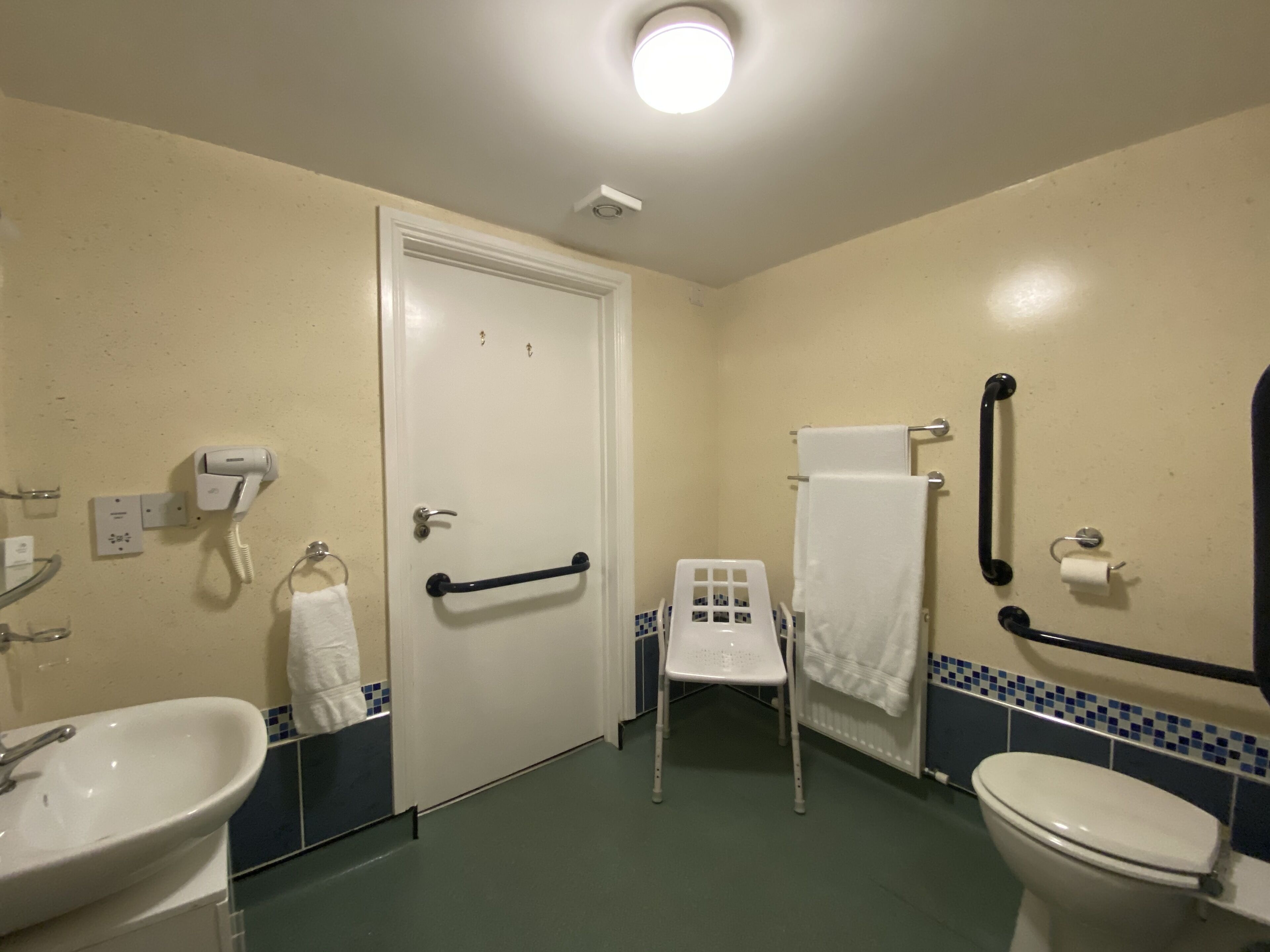 Ein kleines Badezimmer mit Waschbecken, Toilette und Spiegel. Eine weiße Tür mit Griff, ein Handtuchhalter und ein kleiner Stuhl sind ebenfalls vorhanden.