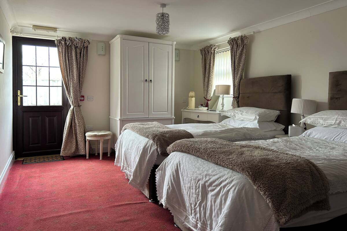 A hotel room with two beds, a wardrobe, a bedside table with a lamp, and a small stool.