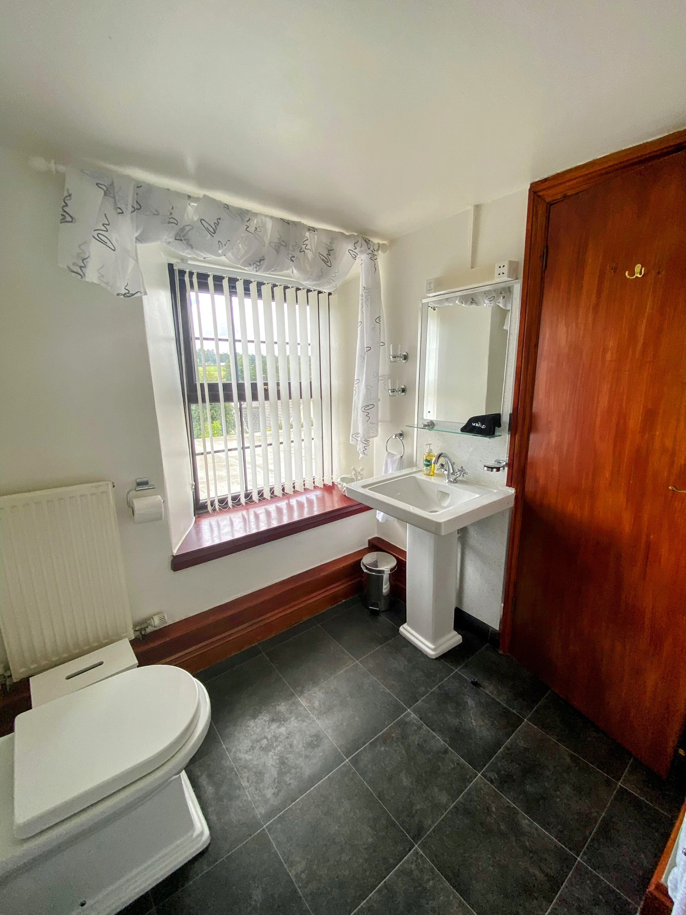 A bathroom with a toilet, sink, and mirror. There is a window with vertical blinds and a curtain. A radiator is on the wall, and a wooden door is visible.